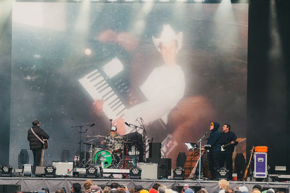 Band performing on outdoor festival stage with large screen displaying keyboard player in white hat.