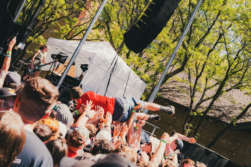 Crowd surfing at an outdoor music festival with lush green trees and a stage in the background.