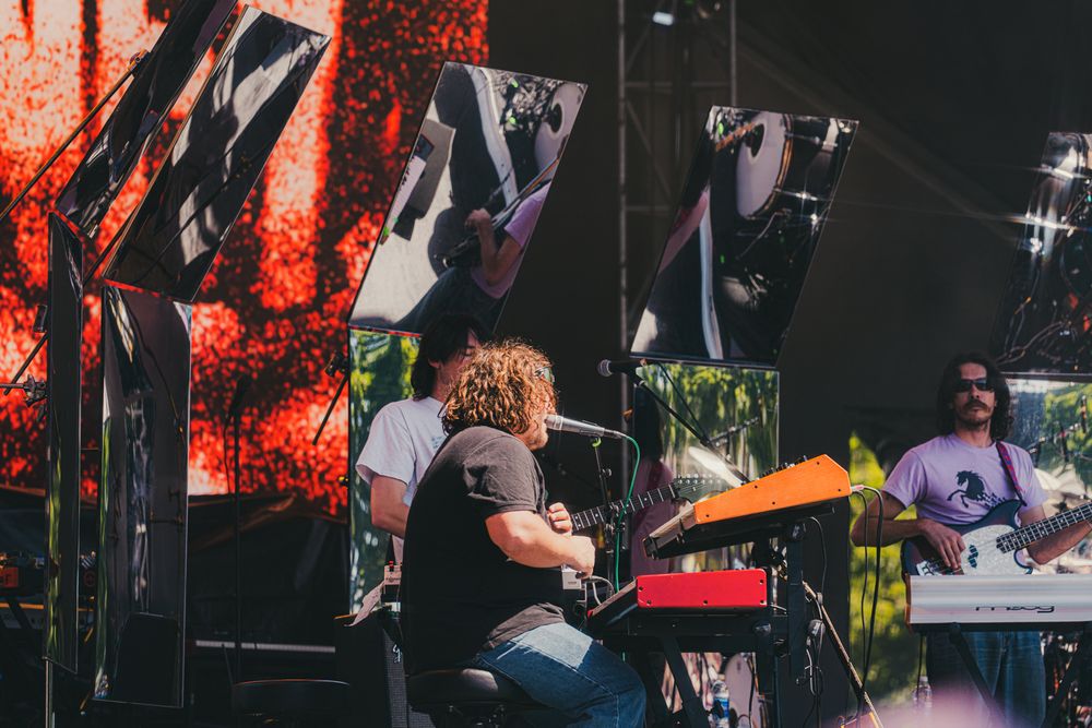 Musicians perform on an outdoor festival stage with red lighting backdrop and speaker arrays.