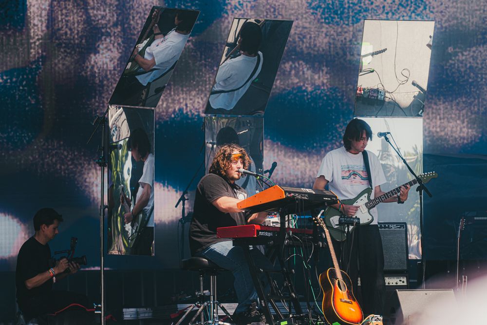 Band performing on stage with guitarist in orange shirt, electric guitars, and large projected screen backdrop at outdoor concert.