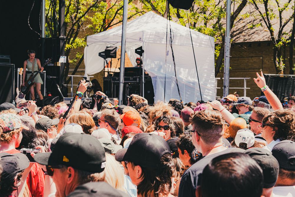 Energetic outdoor music festival crowd cheering in front of a white stage on a sunny day.