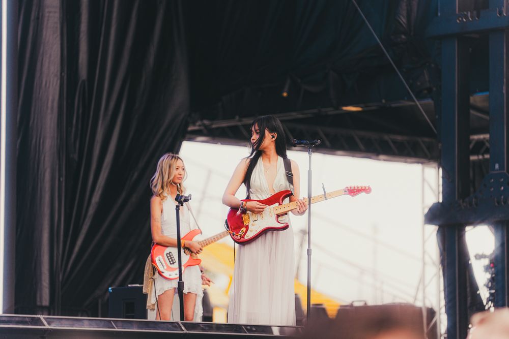 Two women performing on stage, one in white dress playing red electric guitar, other singing at outdoor music festival.
