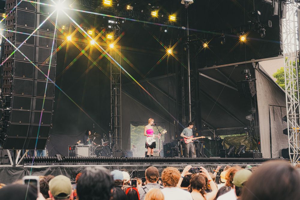 Live outdoor music festival with performers on a bright stage, surrounded by a large crowd under colorful stage lights.