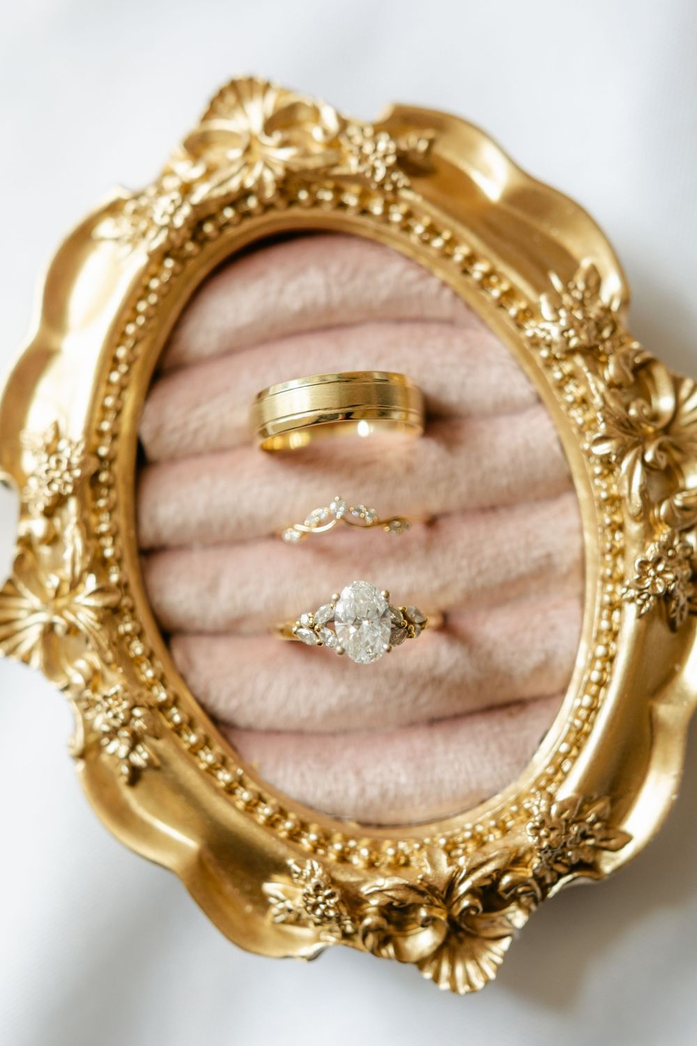 Three gold wedding rings displayed in ornate gold oval frame on pink velvet.
