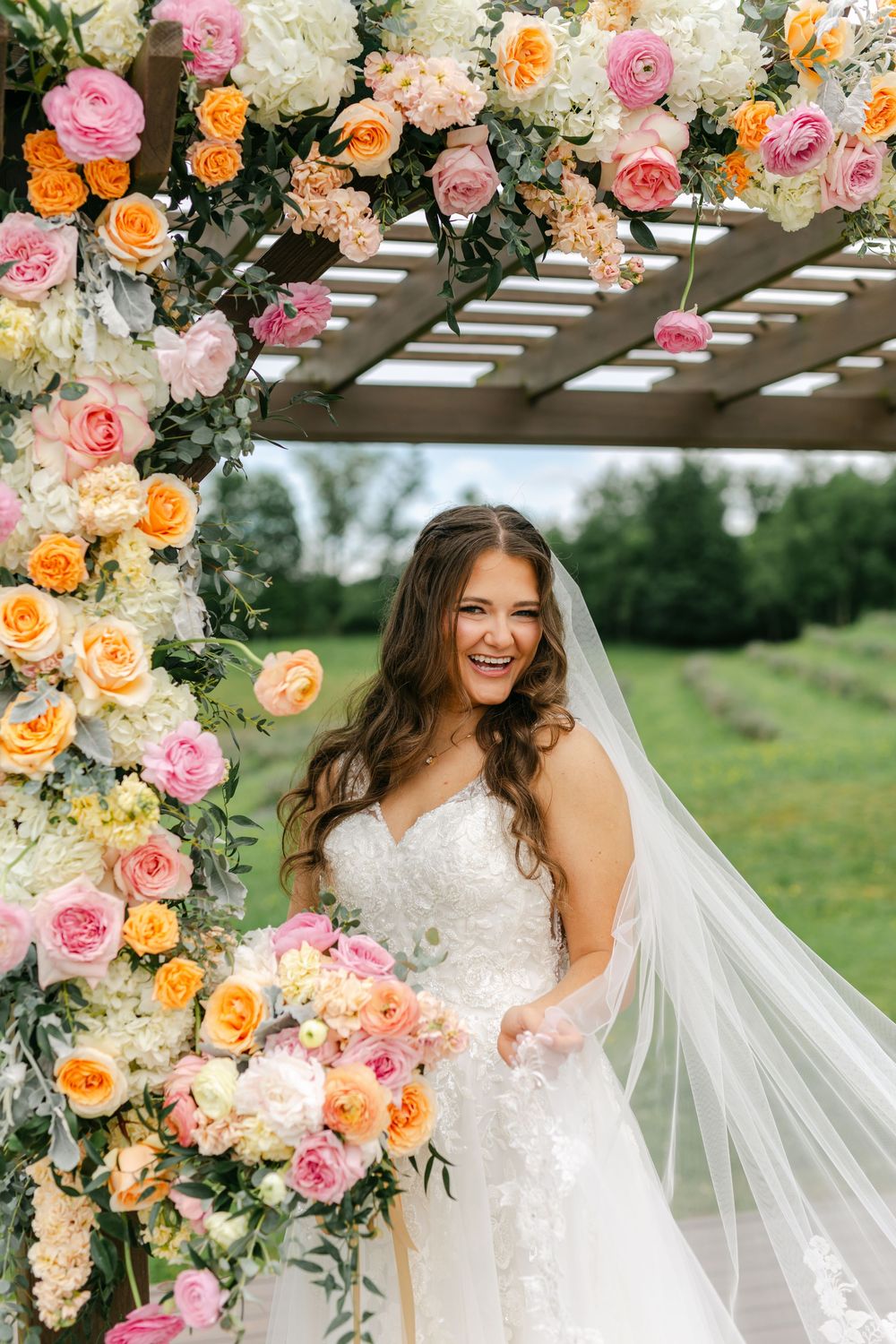 Bride smiling under vibrant pink and orange floral arch holding bouquet with veil flowing.