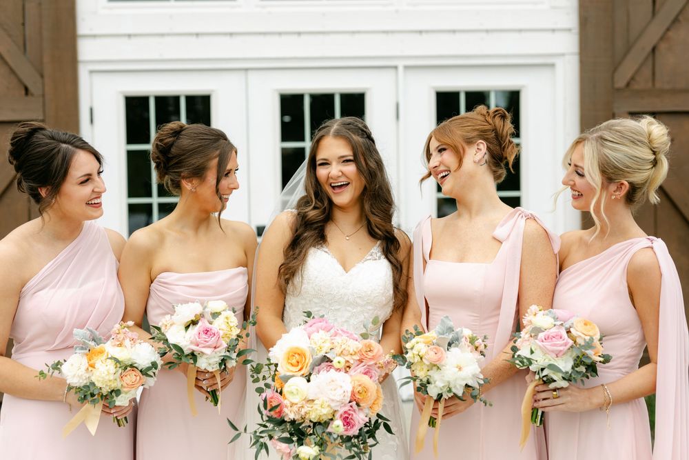 Bride laughs joyfully with four bridesmaids in pink dresses holding colorful bouquets outside barn.