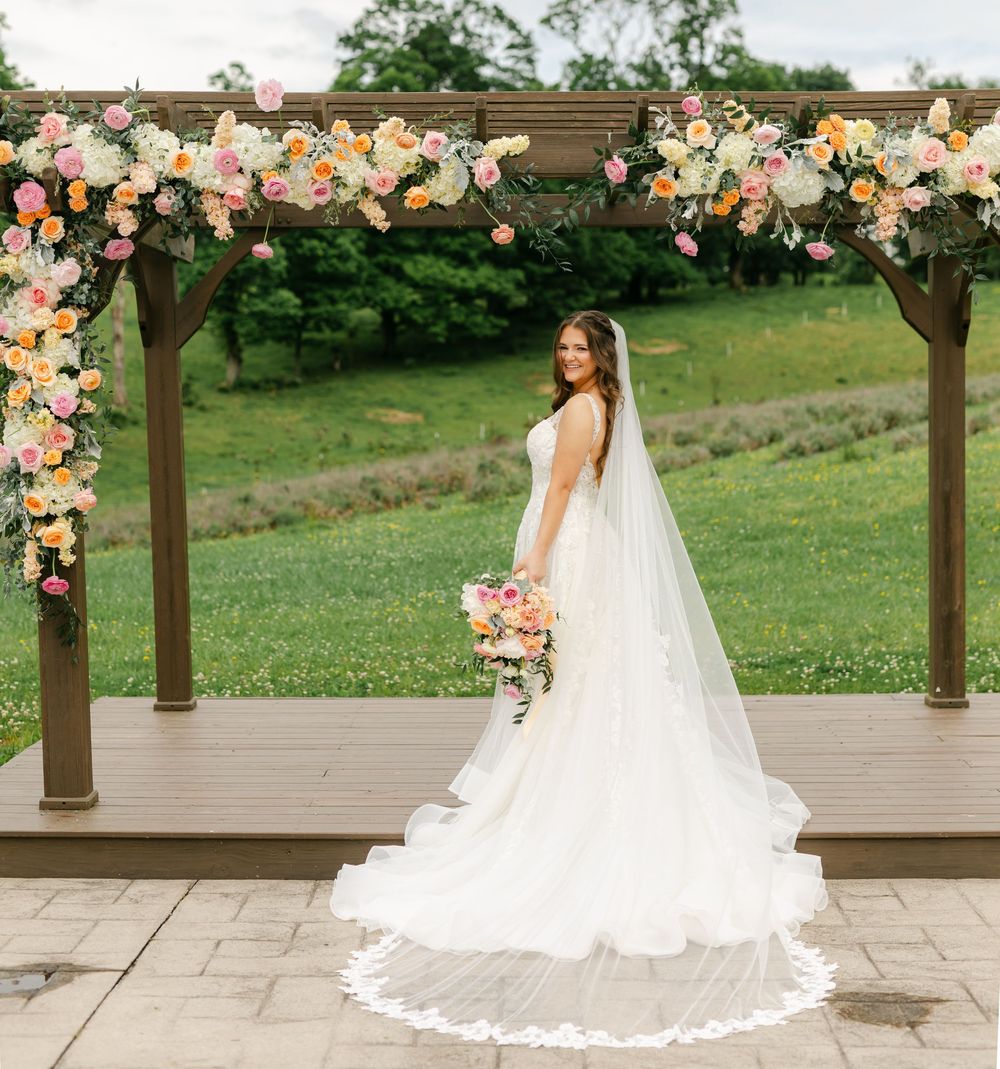Bride in lace gown and veil posing under floral pergola with colorful bouquet outdoors.
