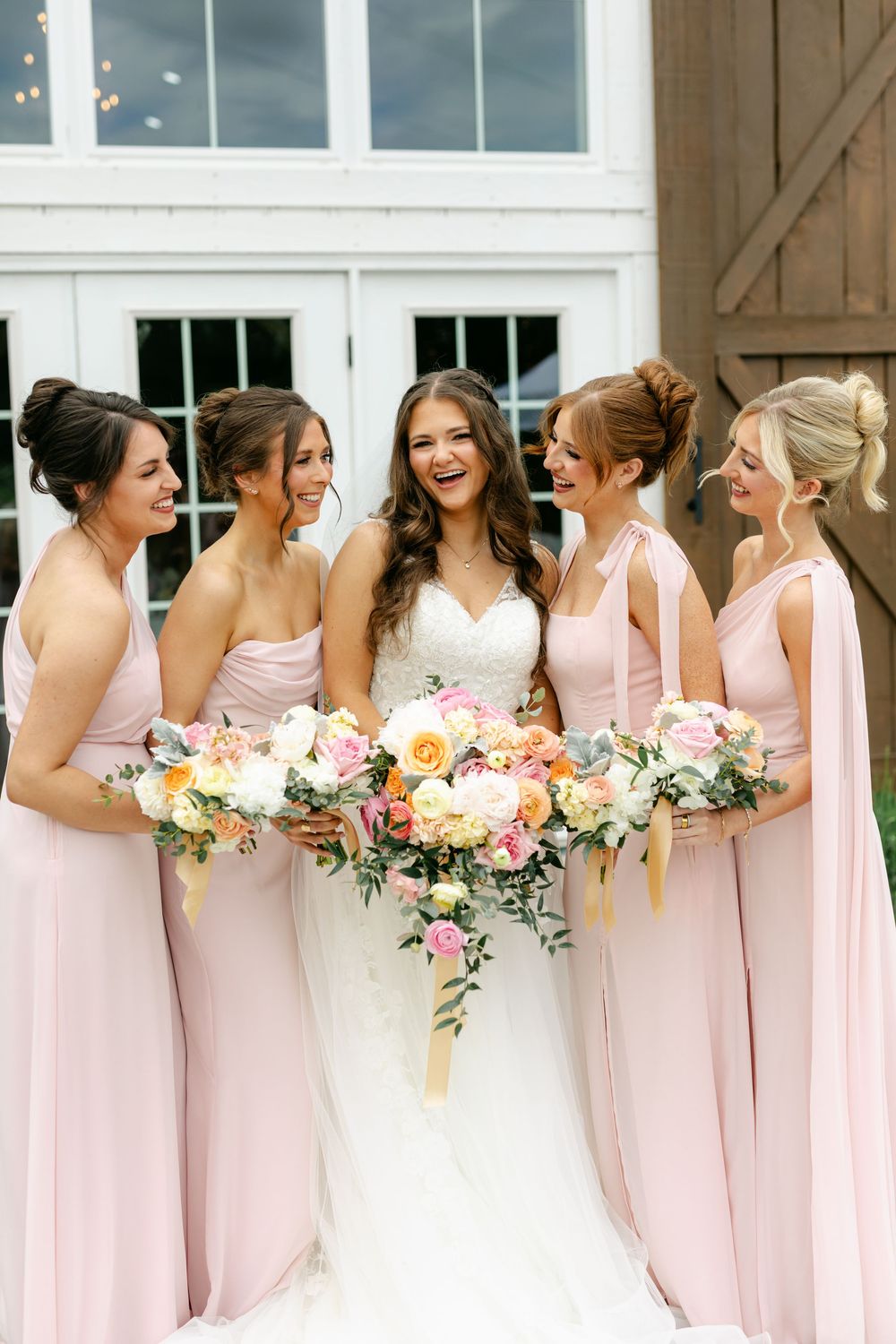 Bride laughing with four bridesmaids in blush pink gowns holding colorful floral bouquets.