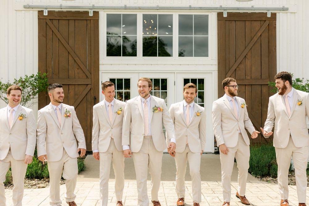 Groomsmen in matching tan suits with boutonnieres posing in front of white barn doors at outdoor wedding.
