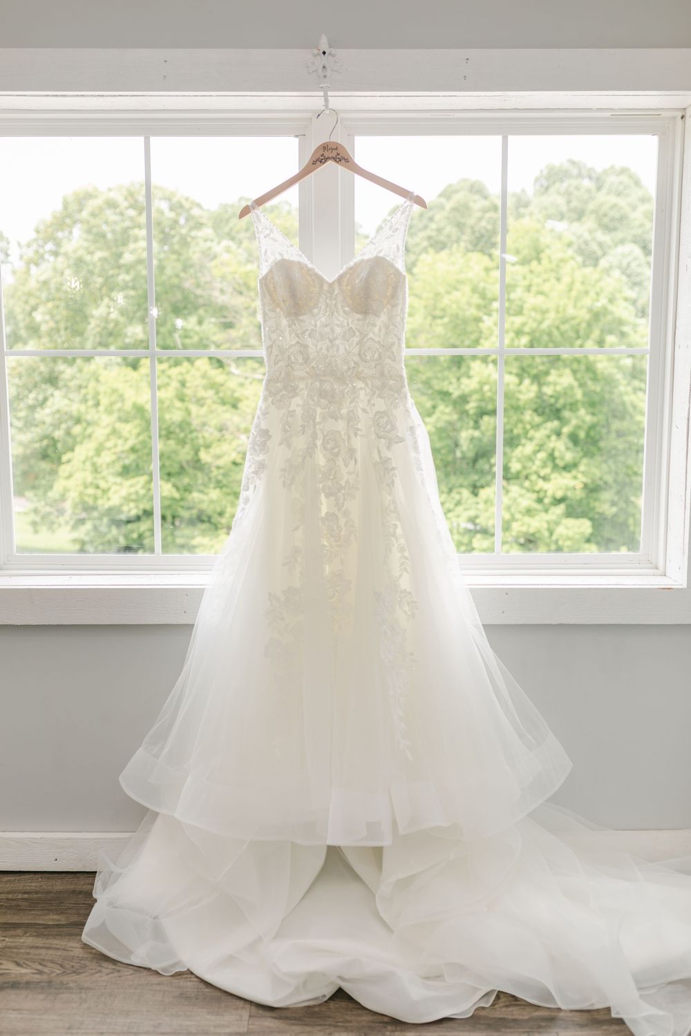 Elegant lace A-line wedding dress hanging in window with green trees in background.