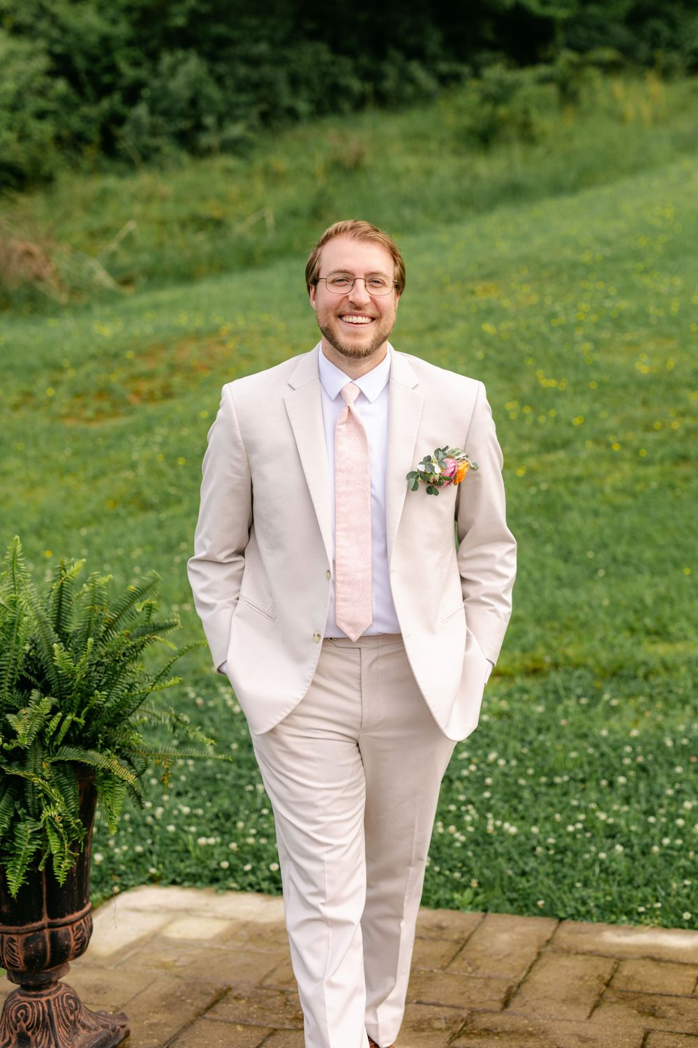 Smiling groom in beige suit with floral boutonniere standing outdoors on stone patio with green hillside backdrop.