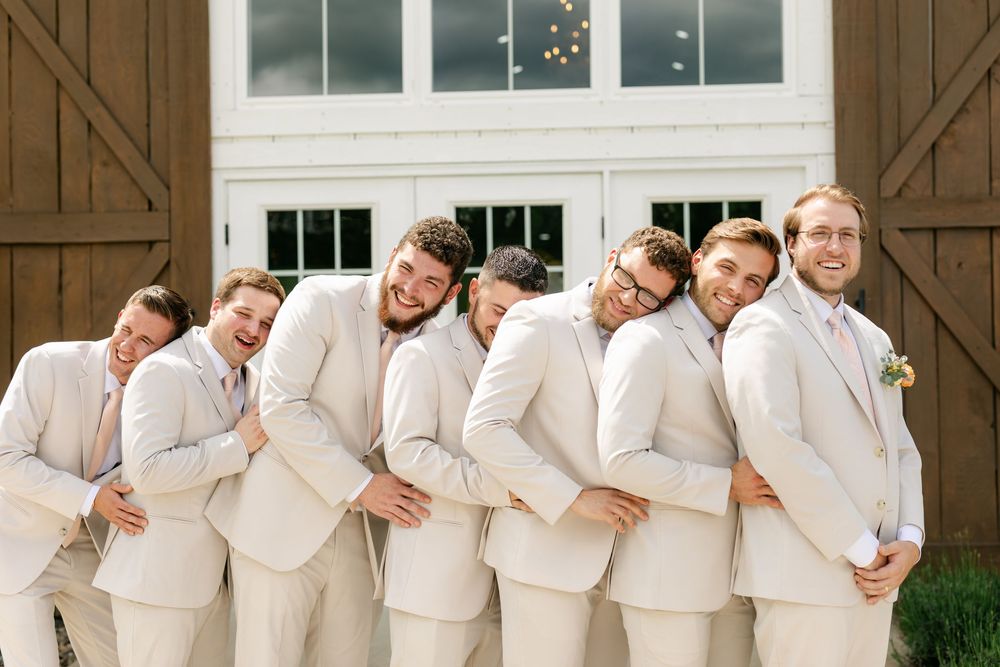 Groomsmen in cream suits playfully posing in a train line in front of rustic barn wedding venue.