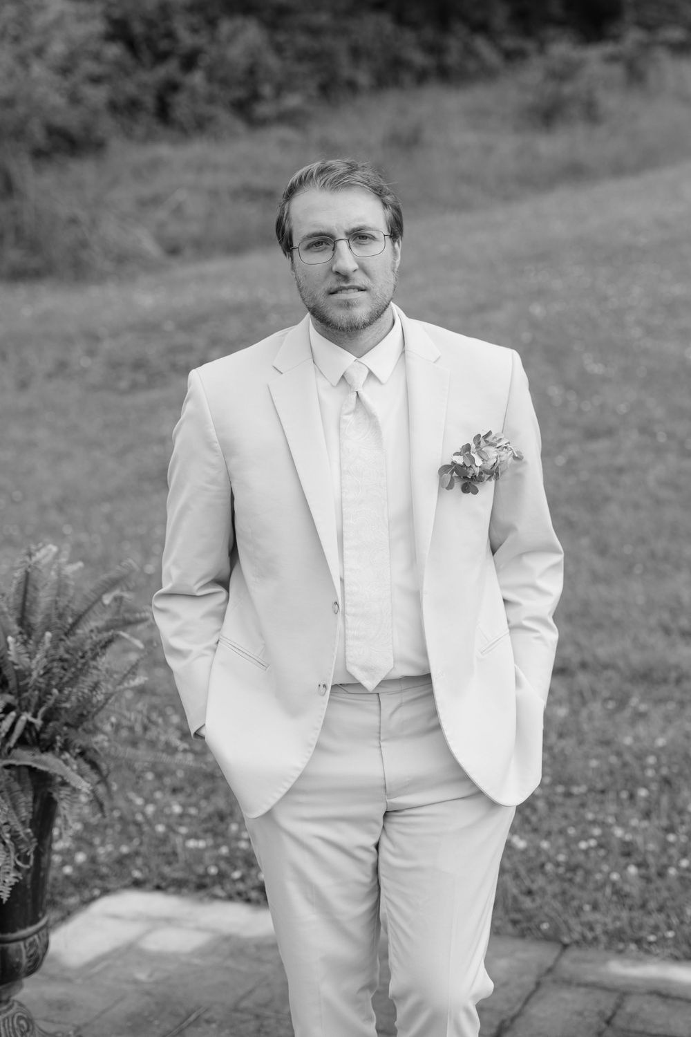 Black and white portrait of groom in light suit with boutonniere standing on stone patio at outdoor wedding.
