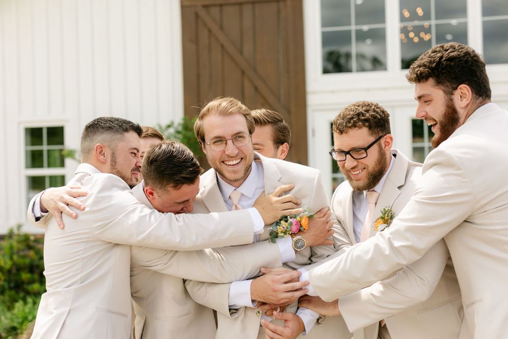 Groomsmen in tan suits sharing a group hug and laughing outside white barn wedding venue.