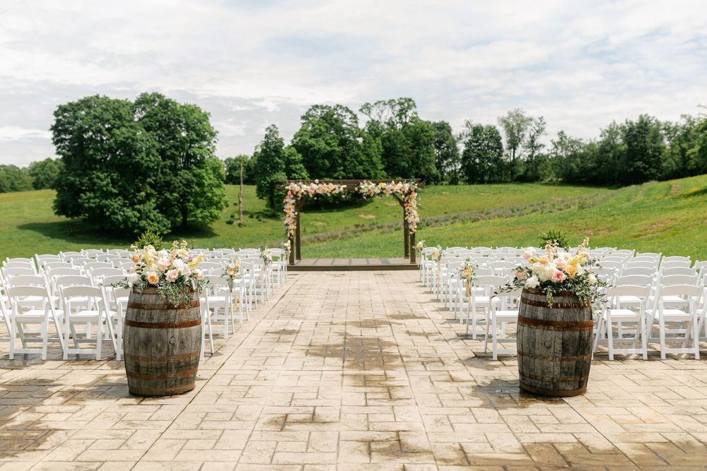 Outdoor wedding ceremony setup with wooden arch, colorful floral arrangements, white chairs, and barrel decorations.