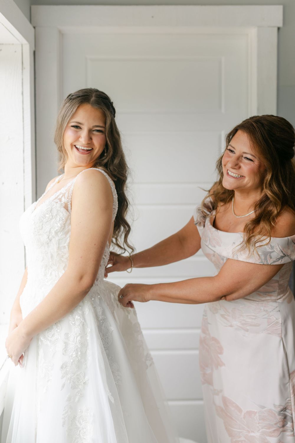 Bride smiling as bridesmaid helps button her white lace wedding gown in a bright white room.
