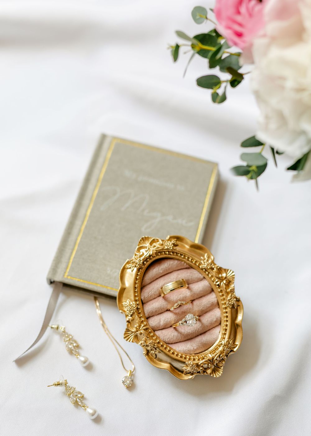 Gold ornate ring dish with wedding bands beside a personalized journal and pearl earrings.