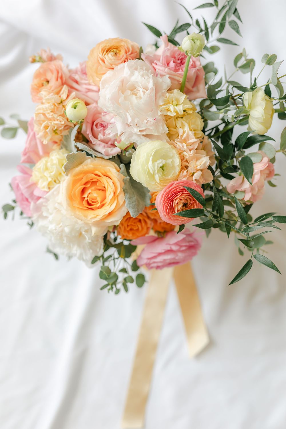 Bridal bouquet of peach, pink, and white roses with eucalyptus and gold ribbon on white fabric.