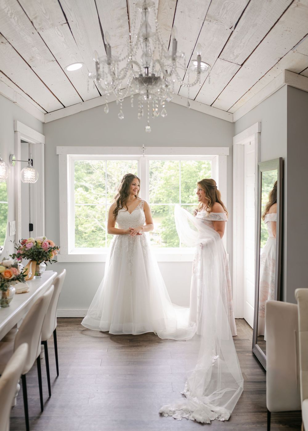 Bride in white gown standing with bridesmaid in elegant bridal suite with crystal chandelier.