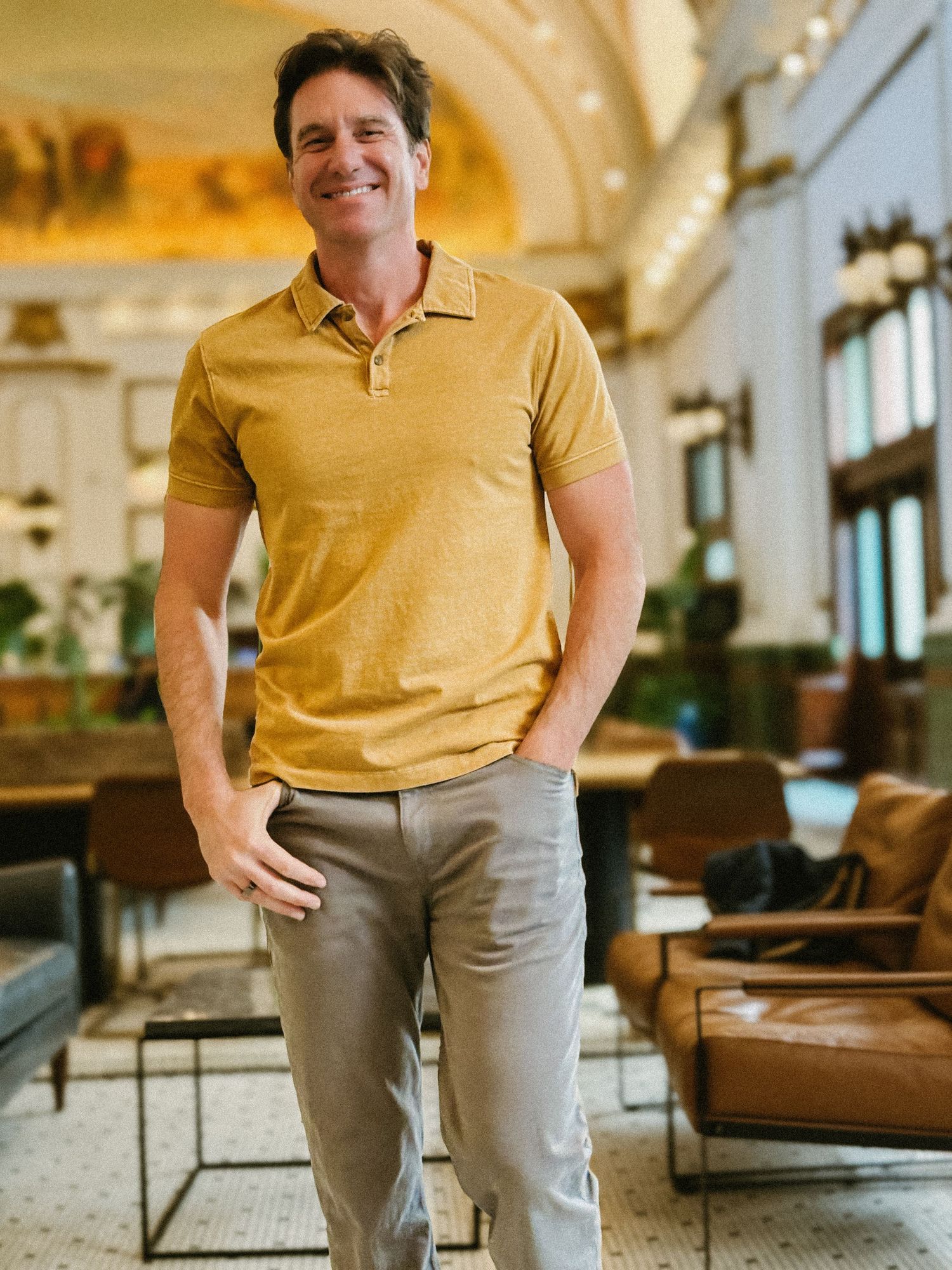 Smiling man in mustard polo shirt standing with hands in pockets in upscale hotel lobby.