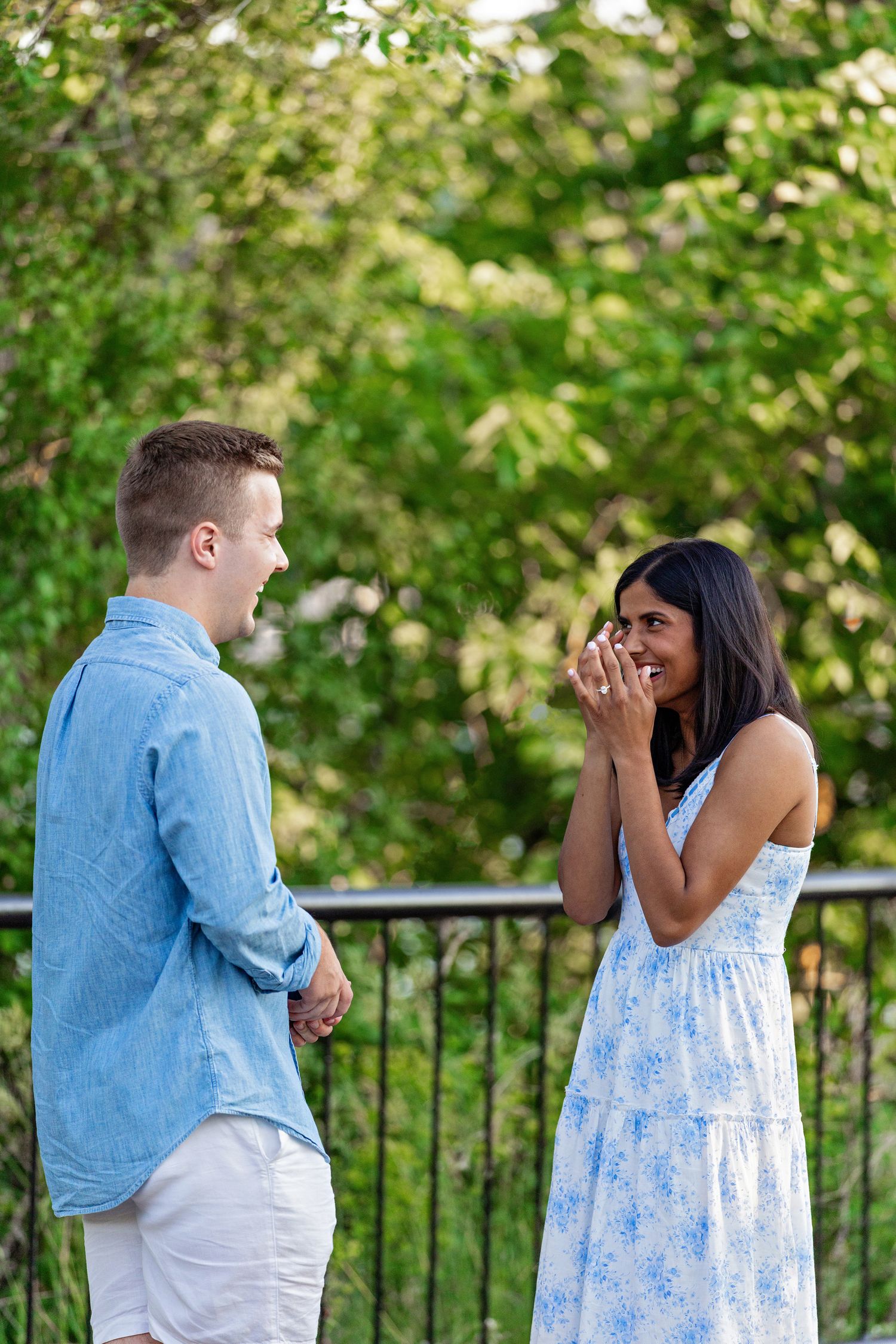 Emotional woman covers her mouth in surprise while man stands beside her after a marriage proposal outdoors.