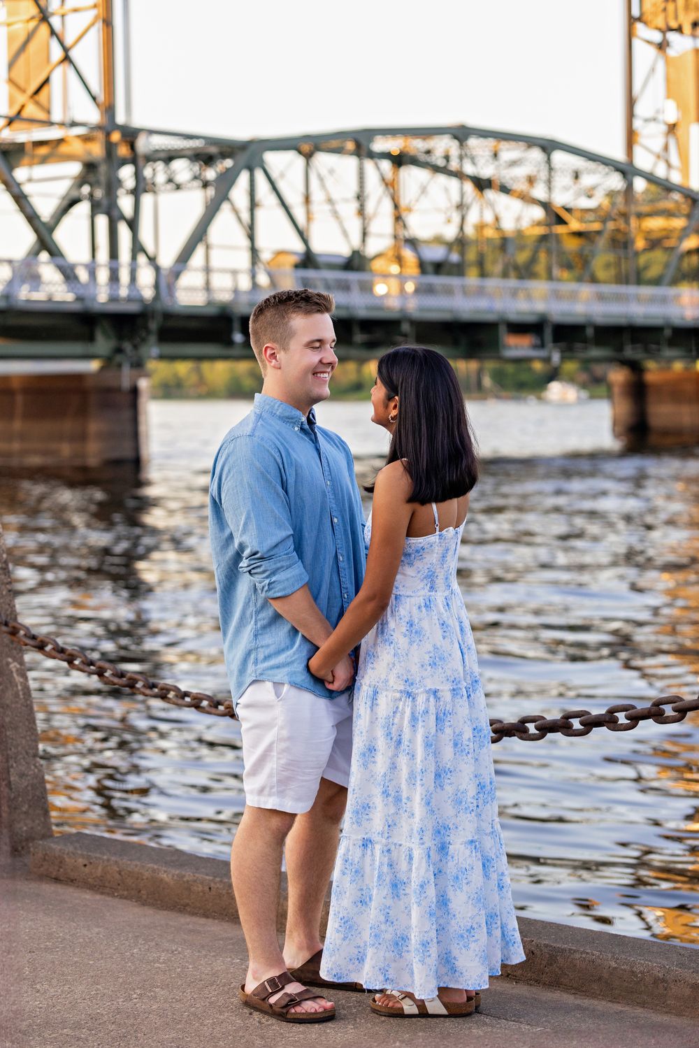Couple holding hands and gazing at each other on a riverside walkway with a bridge view.