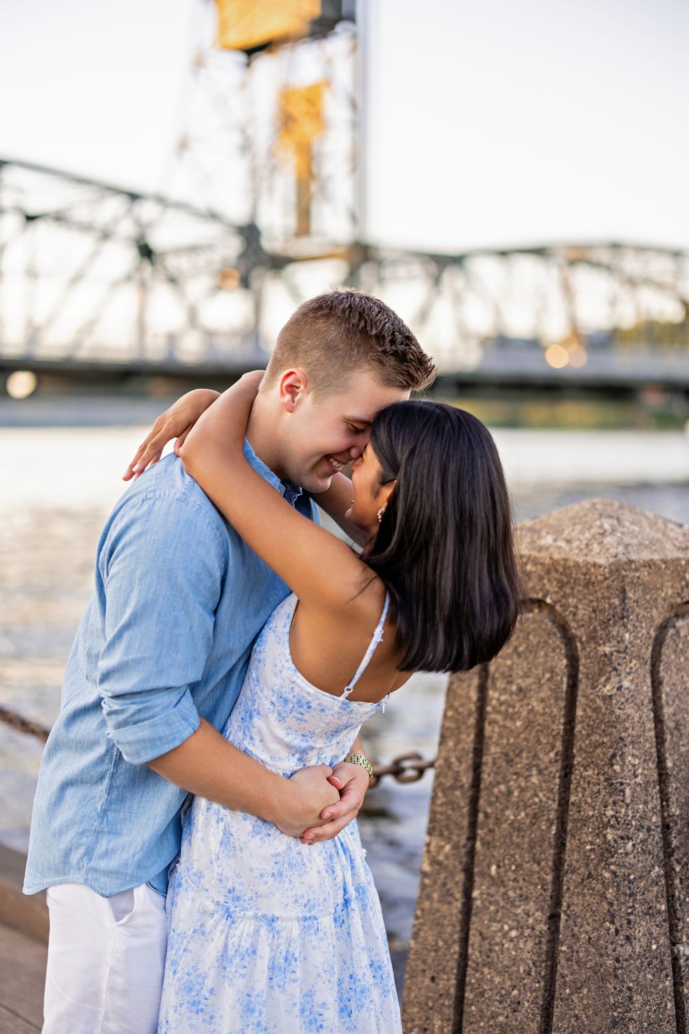 Couple sharing an intimate embrace near a stone bollard by the river with a bridge backdrop.