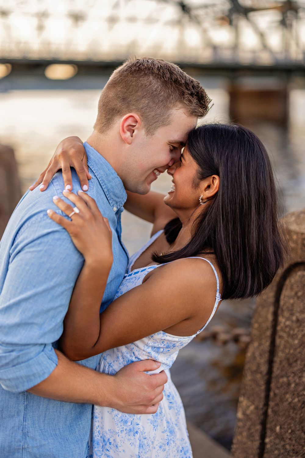 Couple sharing a romantic nose-to-nose moment while embracing near a river with a bridge behind.