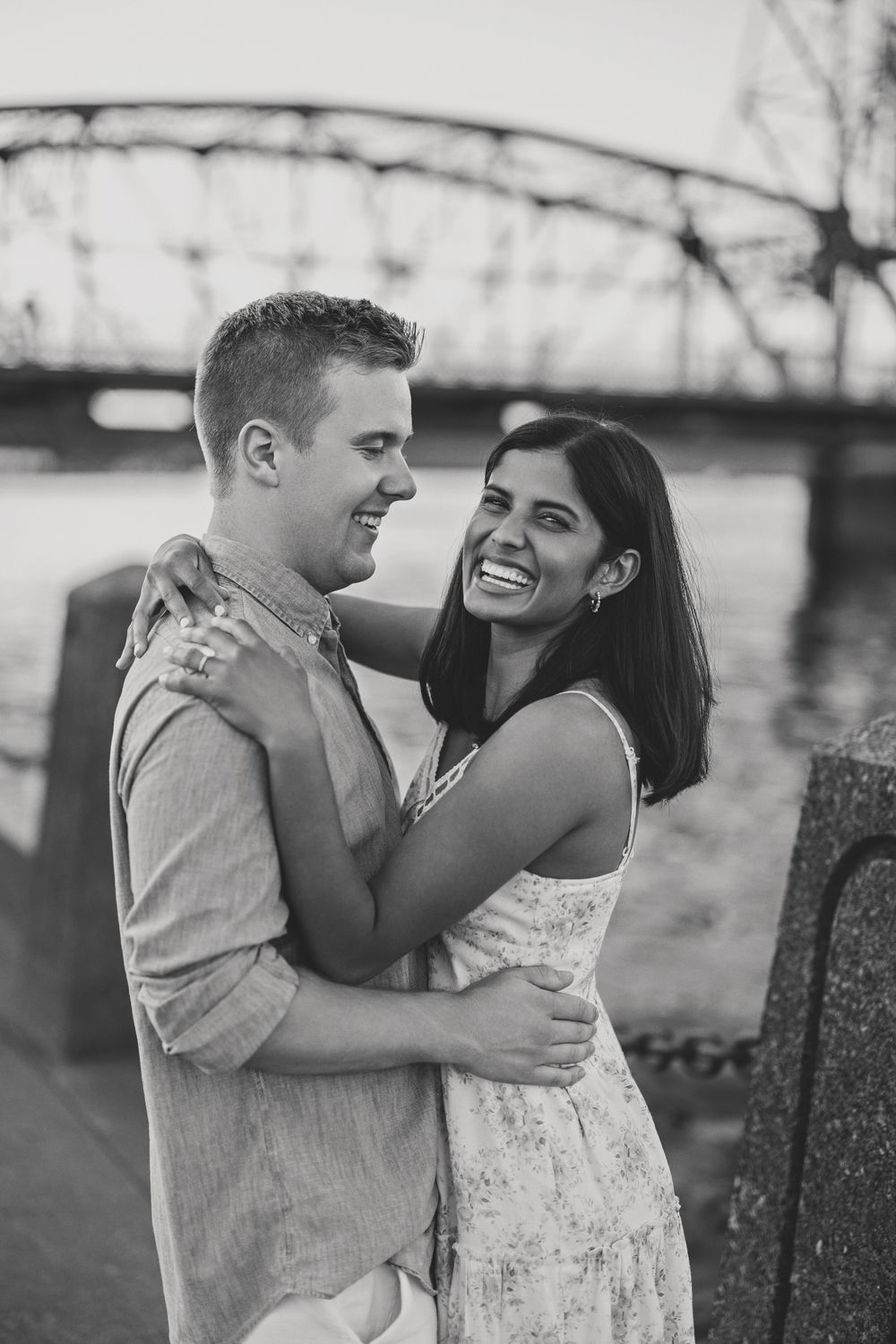Black and white photo of a laughing couple embracing near a river with a steel bridge backdrop.