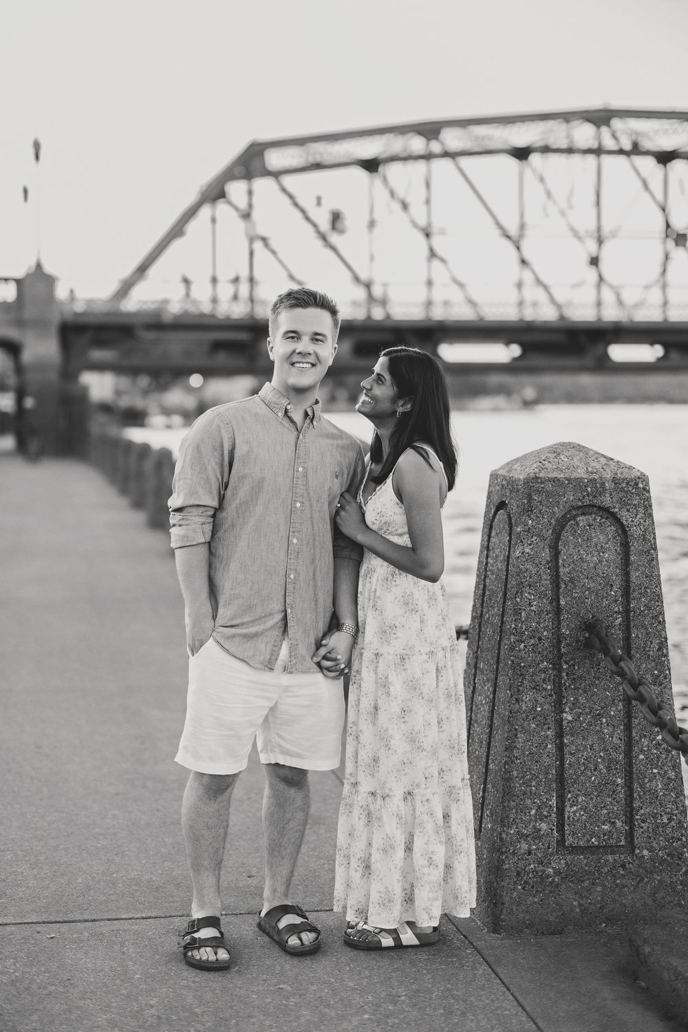 Black and white photo of a couple holding hands on a riverside promenade with a bridge behind.