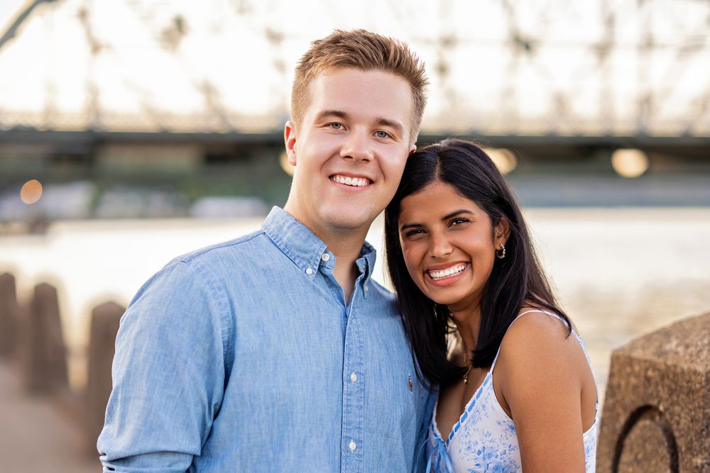 Close-up portrait of a smiling couple posing together near a river with a bridge in background.