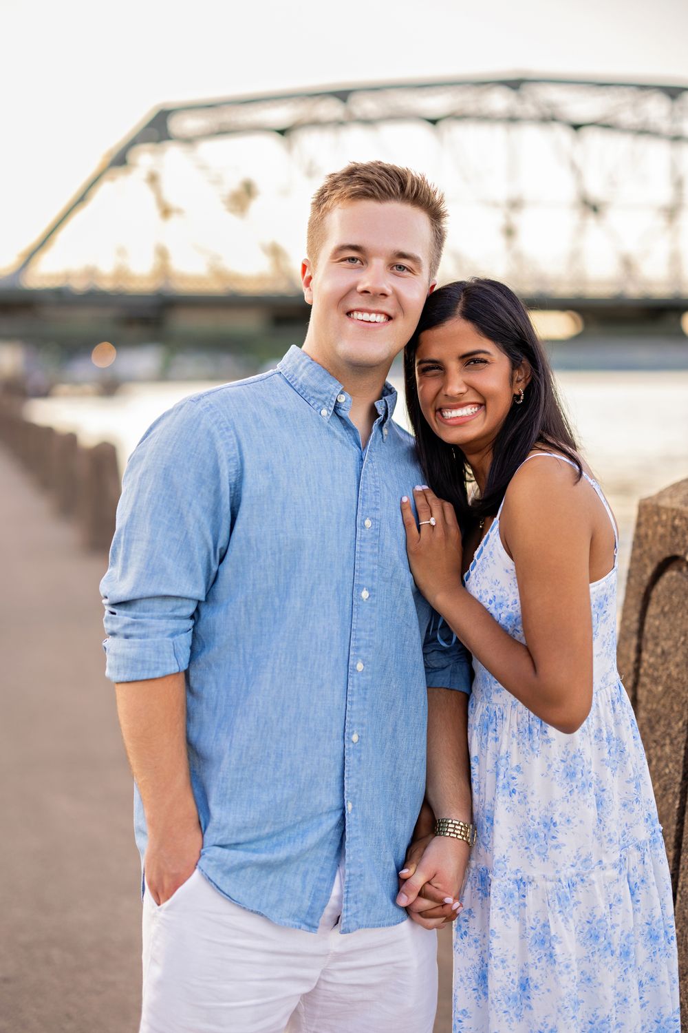Couple smiling and posing together on a waterfront walkway with a steel bridge in the background in Stillwater, MN.