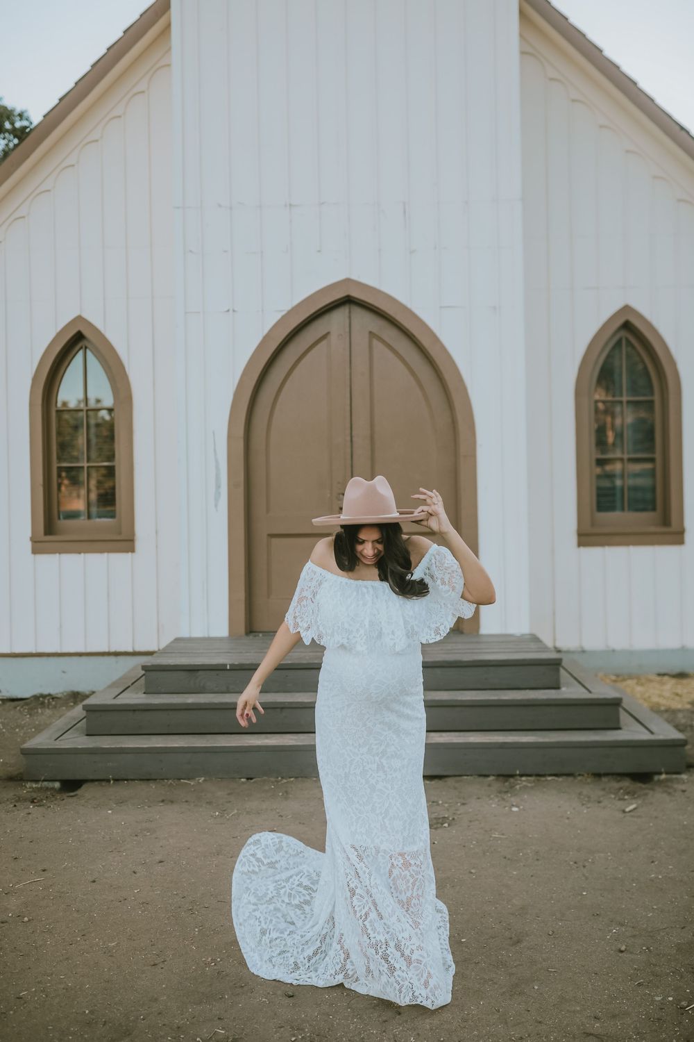 Bride in white lace off-shoulder gown and pink hat poses in front of a white chapel with arched windows.