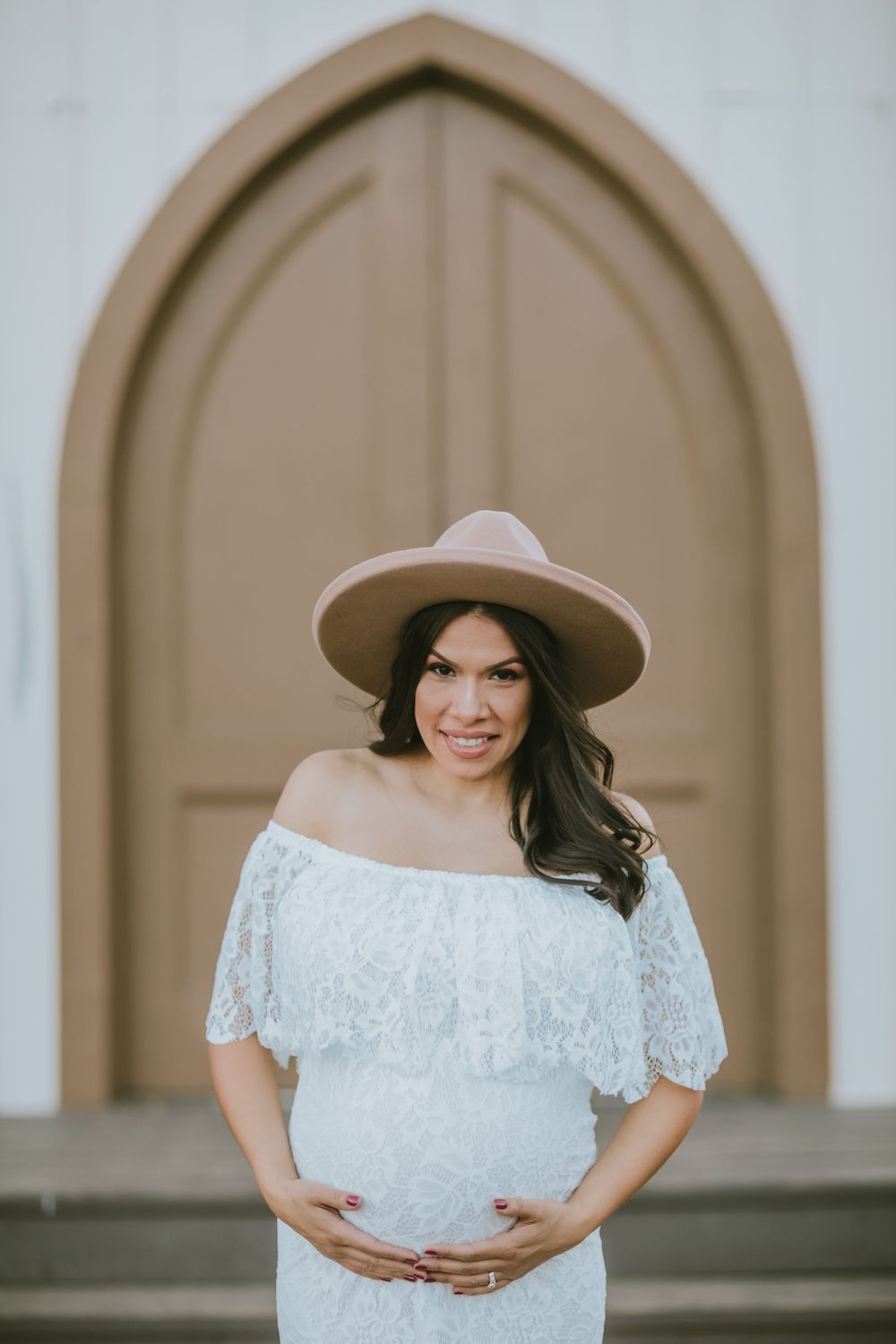 Pregnant woman in white lace off-shoulder dress and beige wide-brim hat poses in front of arched wooden door.