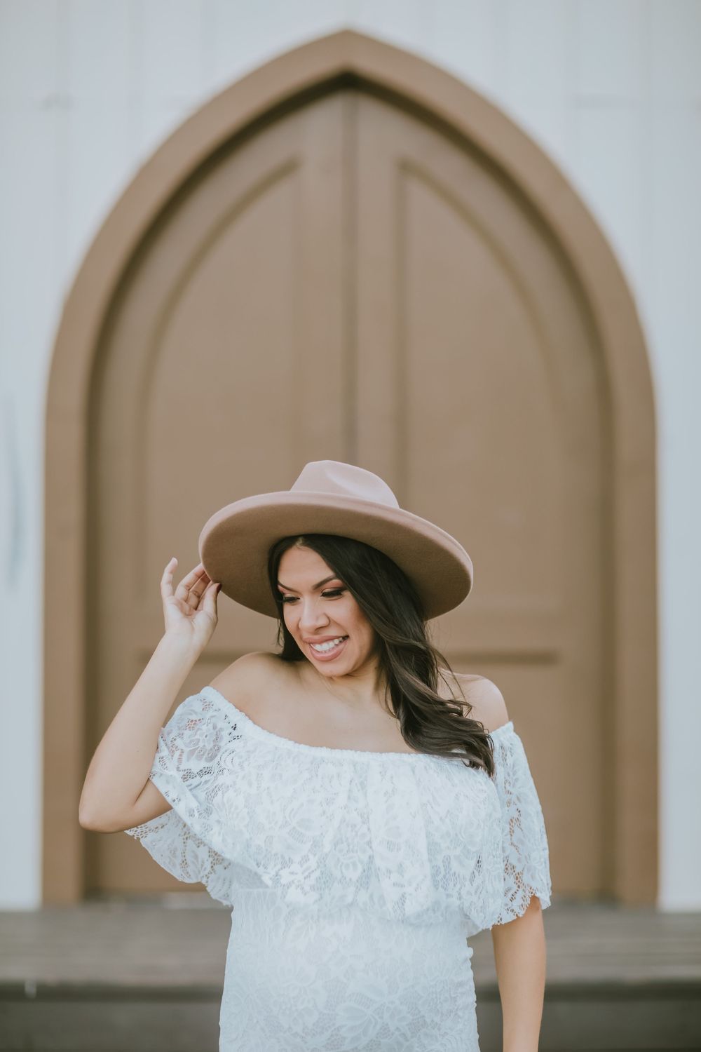 Woman in white off-shoulder lace dress and tan wide-brim hat smiling in front of arched wooden church door.