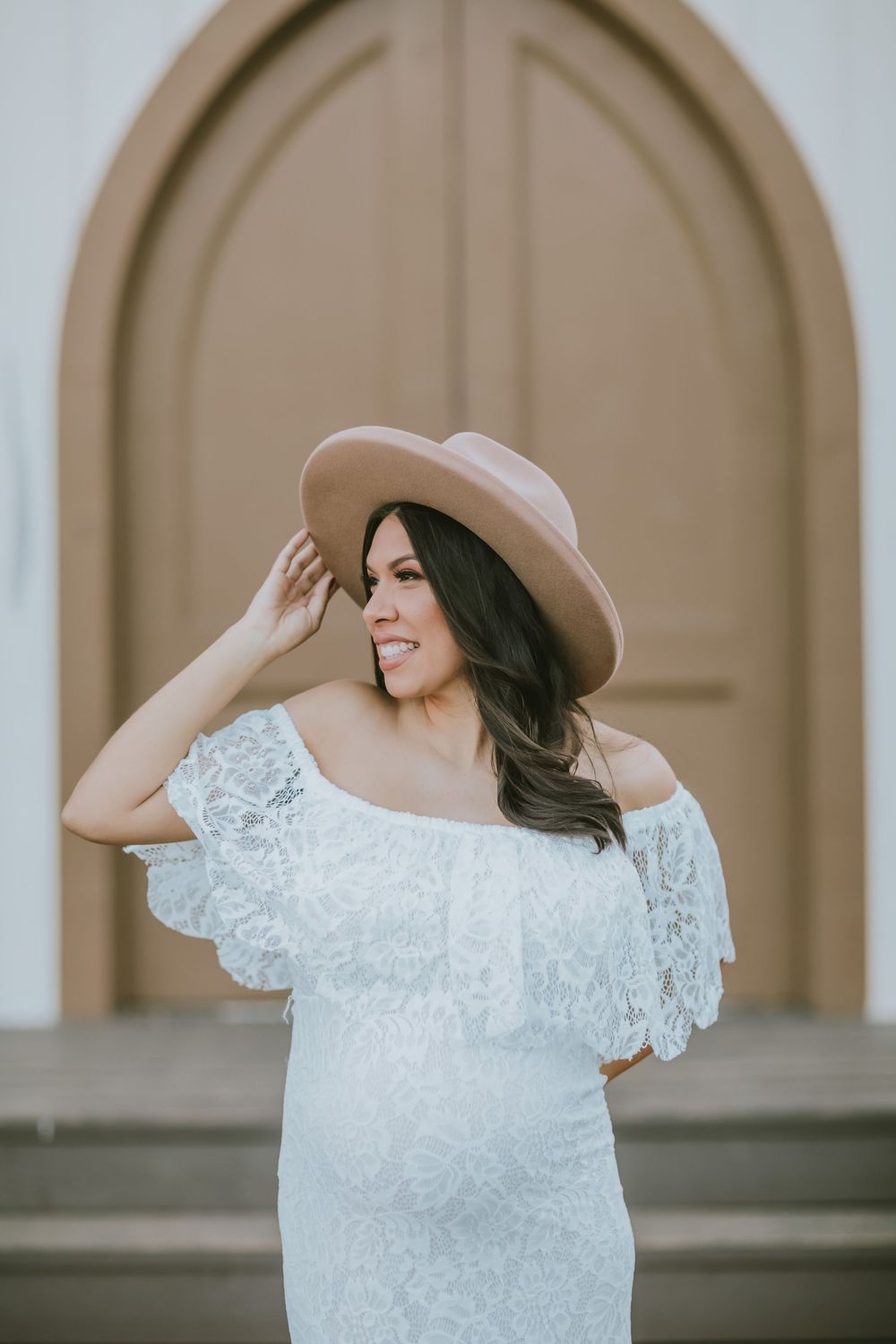 Woman in white off-shoulder lace dress and tan wide-brim hat posing in front of a brown arched wooden door.