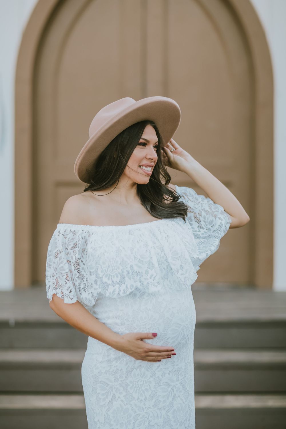 Smiling pregnant woman in white lace off-shoulder dress and beige wide-brim hat poses outdoors near ornate door.