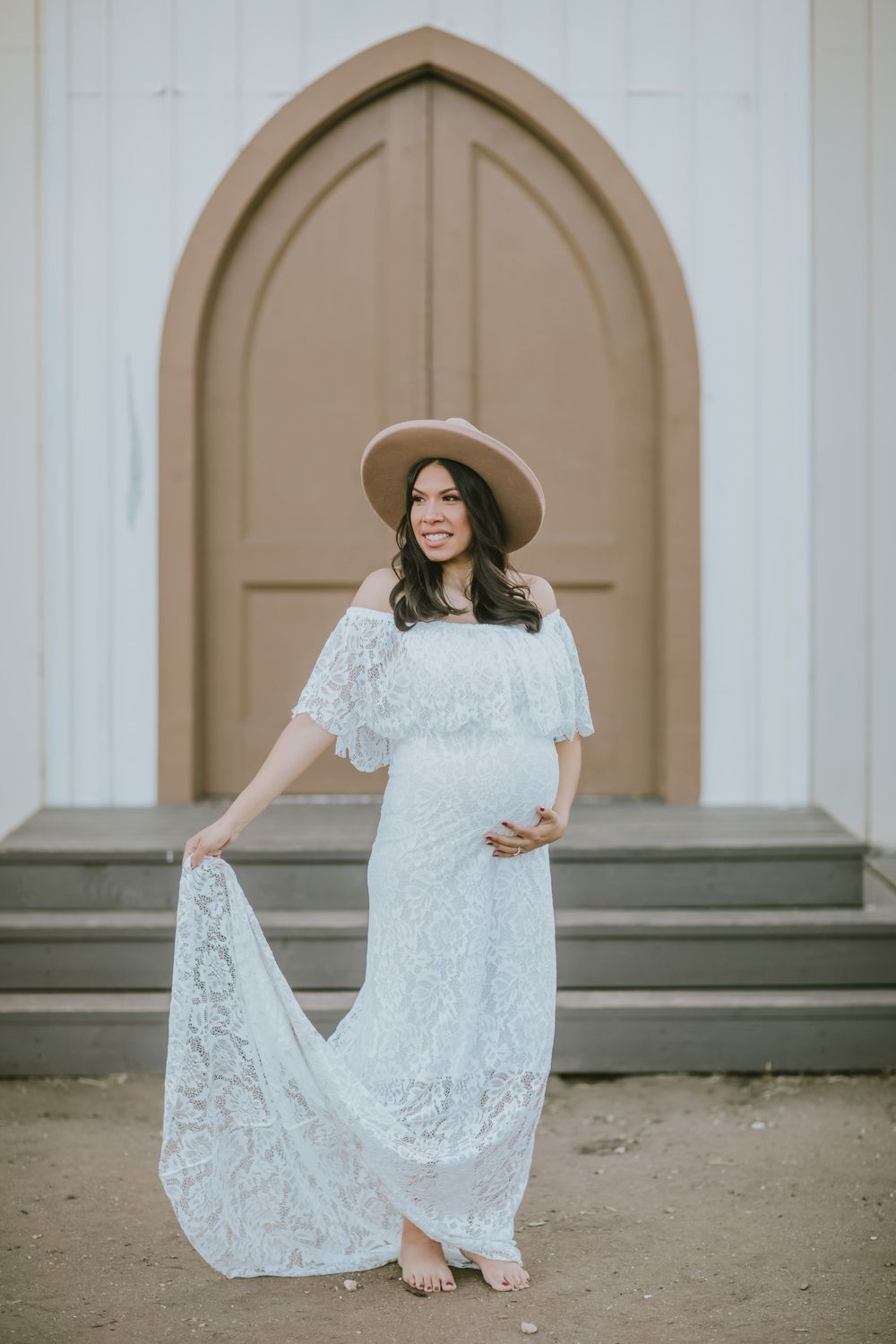 Pregnant woman in white lace off-shoulder gown and tan wide-brim hat poses barefoot before arched church doors.