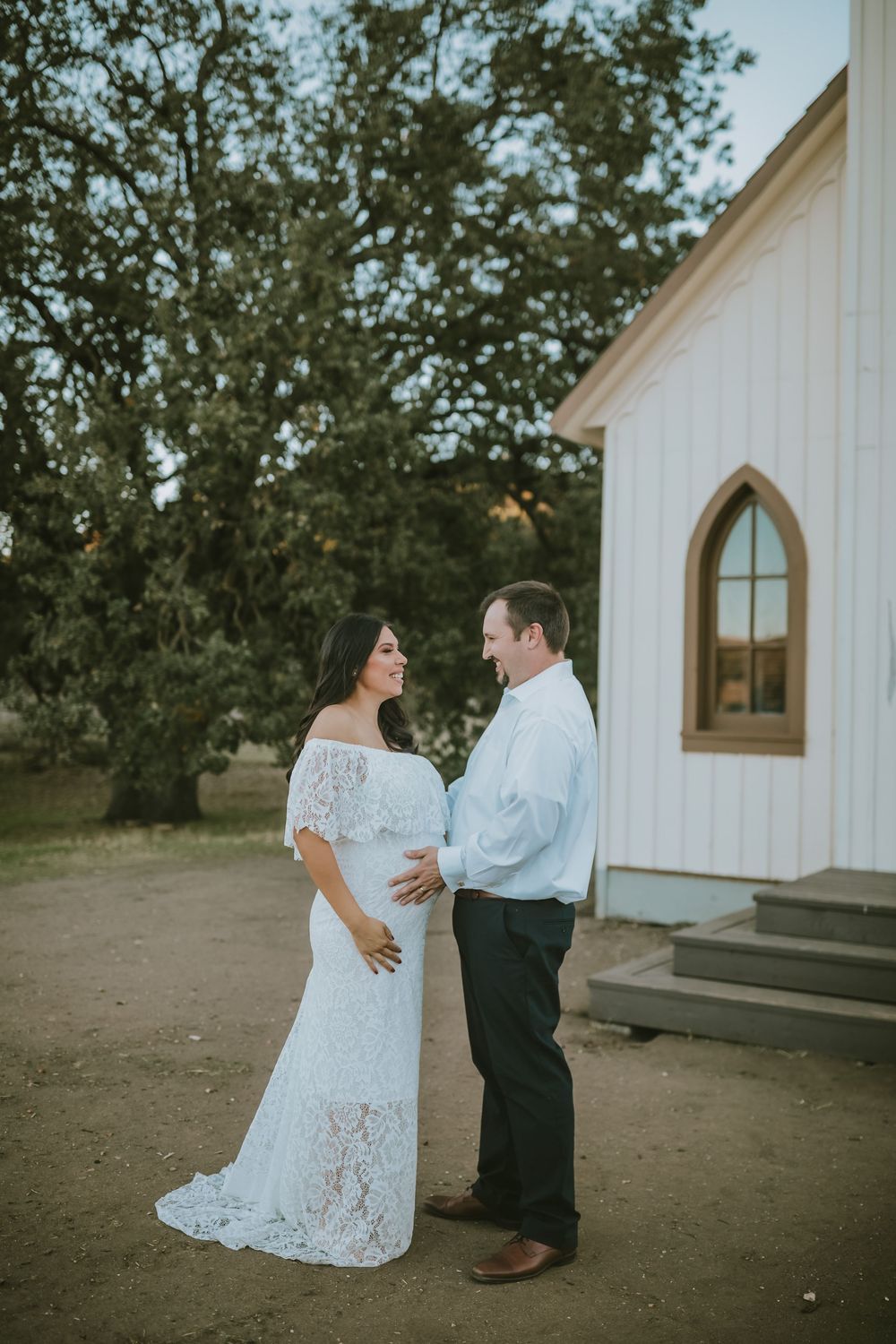 Couple in white lace maternity dress and dress shirt pose lovingly outside a white chapel with arched windows.