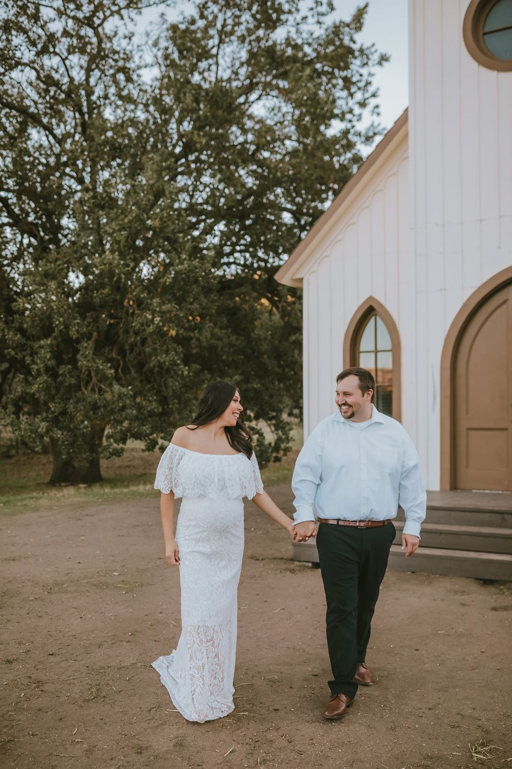 Couple holding hands walking near white chapel, woman in lace off-shoulder gown, man in light blue shirt.