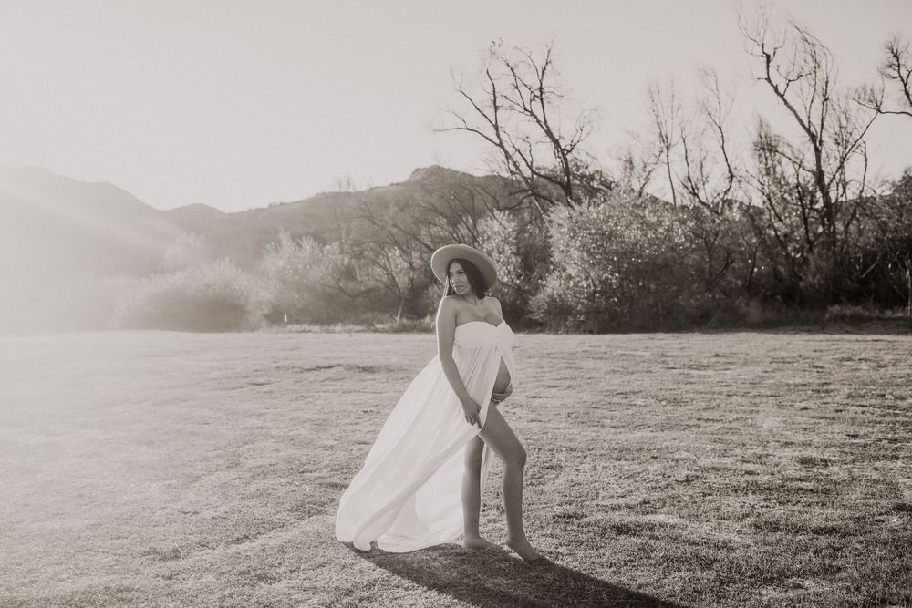 Pregnant woman in white flowing dress and hat poses gracefully in sunlit field with mountain backdrop.