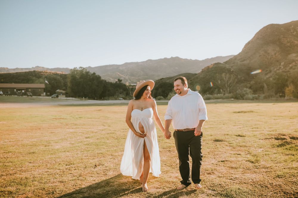 Pregnant couple holding hands walking in sunny field with mountain backdrop, wearing white outfits.