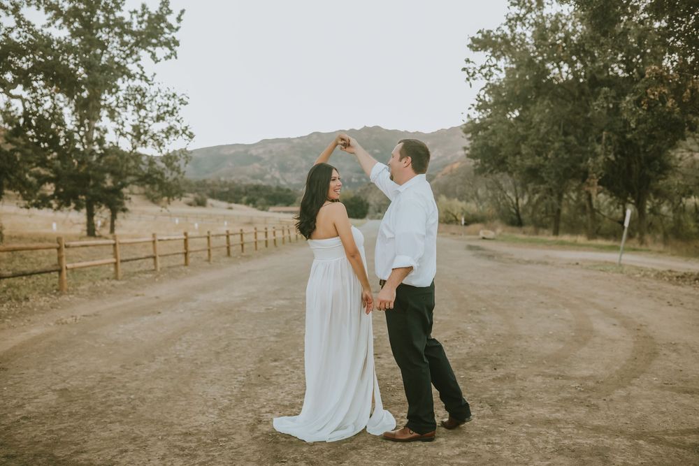 Couple dancing on a dirt road surrounded by trees and hills during golden hour in a romantic outdoor engagement photo.