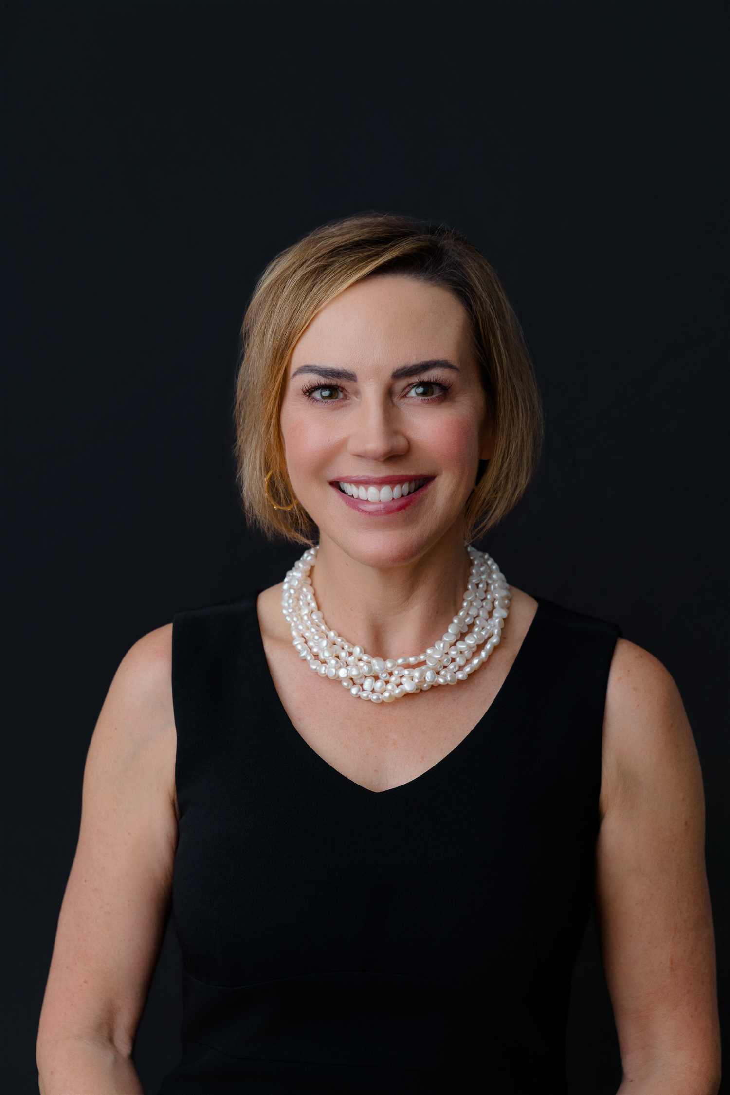 Professional woman with bob haircut wearing black sleeveless top and pearl necklace smiling against dark background.