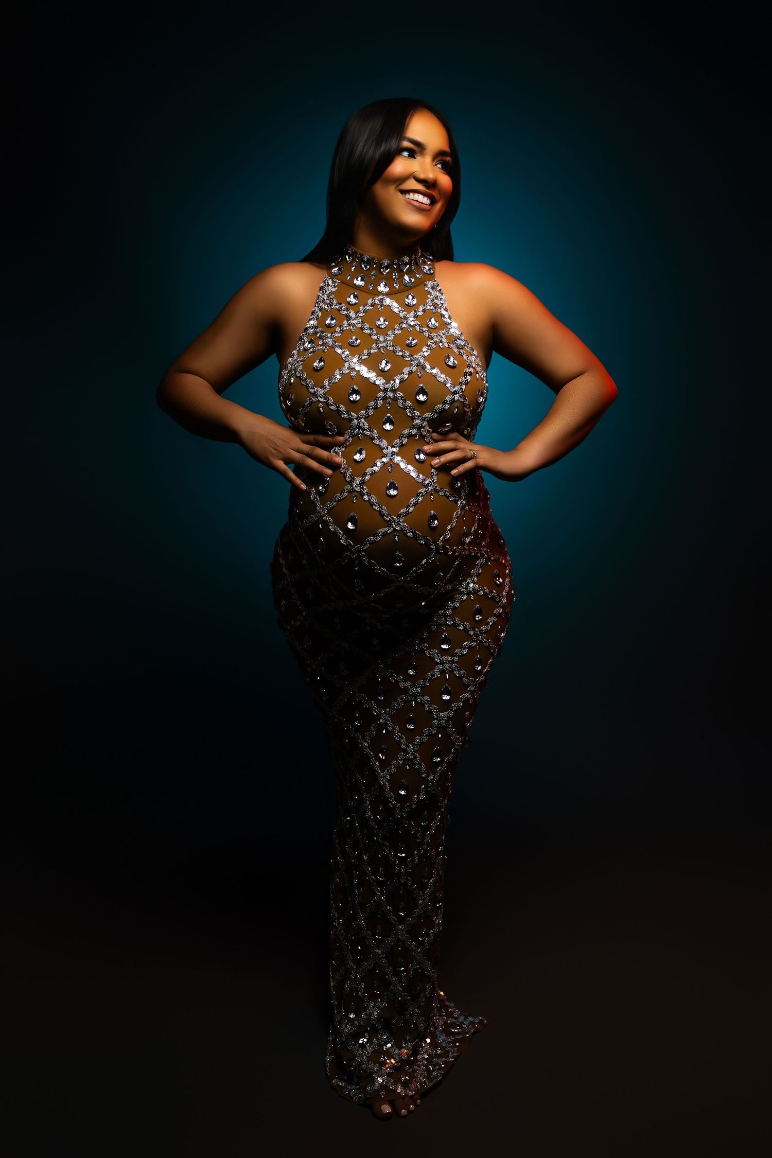 Smiling pregnant woman in sparkling beaded gown posing against teal studio backdrop.