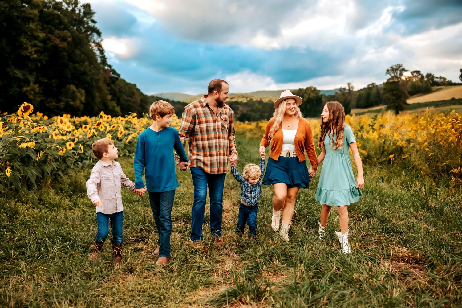 Family of six walking through sunflower field on cloudy day, parents holding hands with children in colorful autumn outfits.