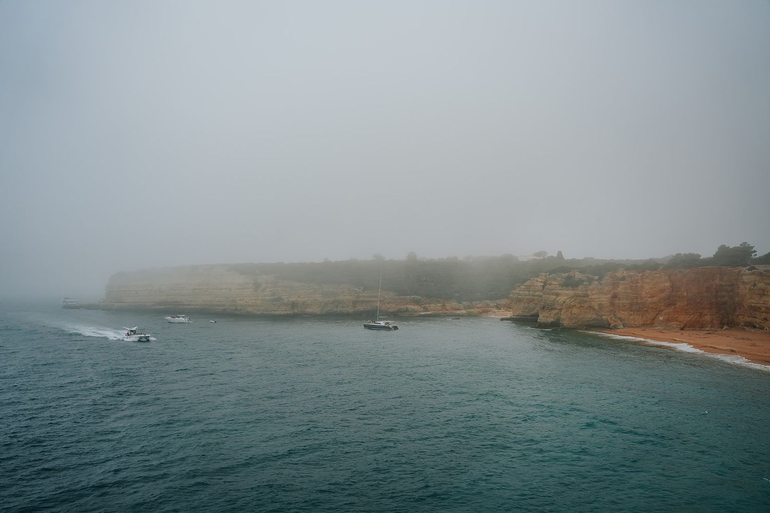 Misty coastal landscape with boats anchored near rugged cliffs and gray waters.
