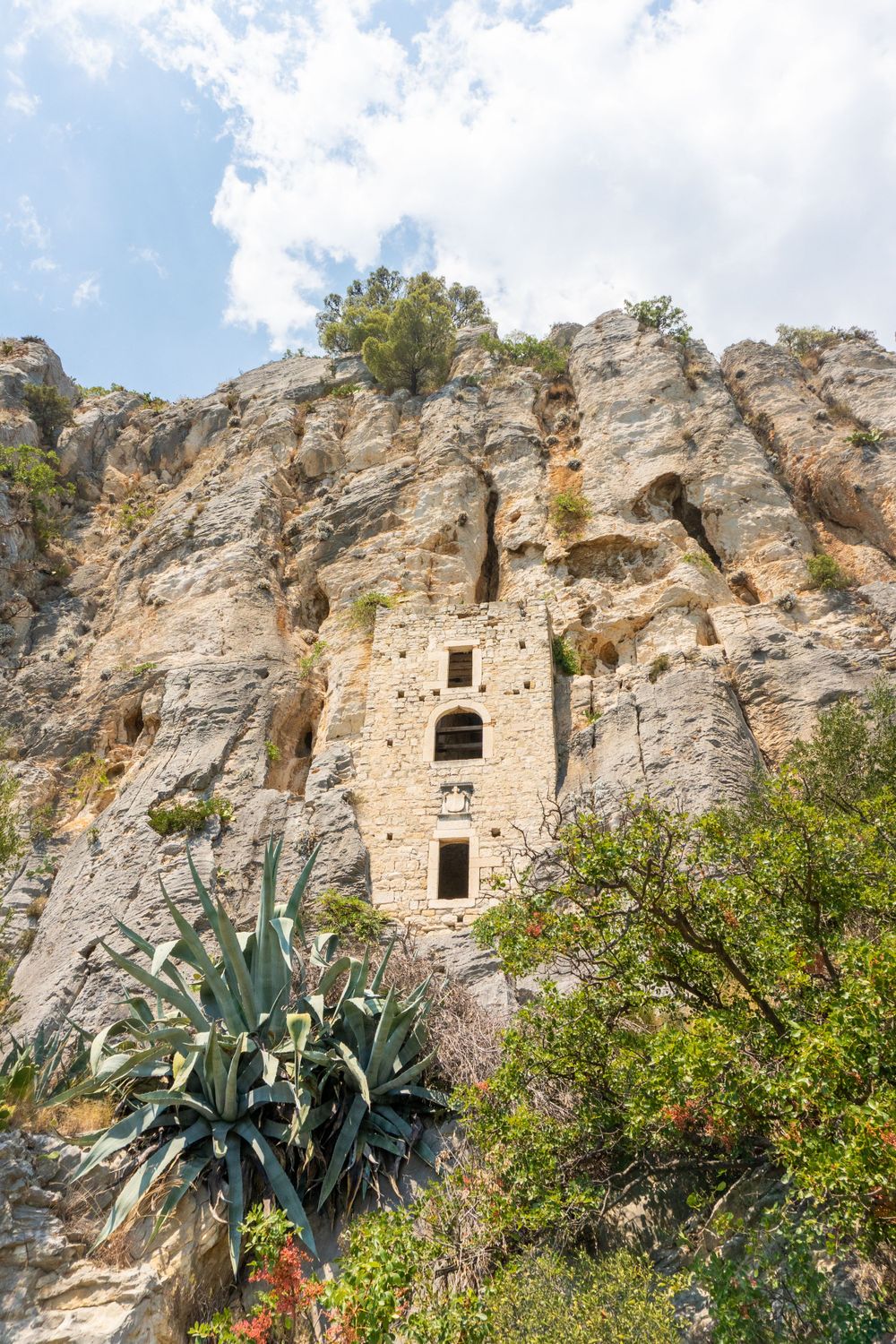 Ancient hermitage monastery built into steep limestone cliffs with agave plants in foreground.