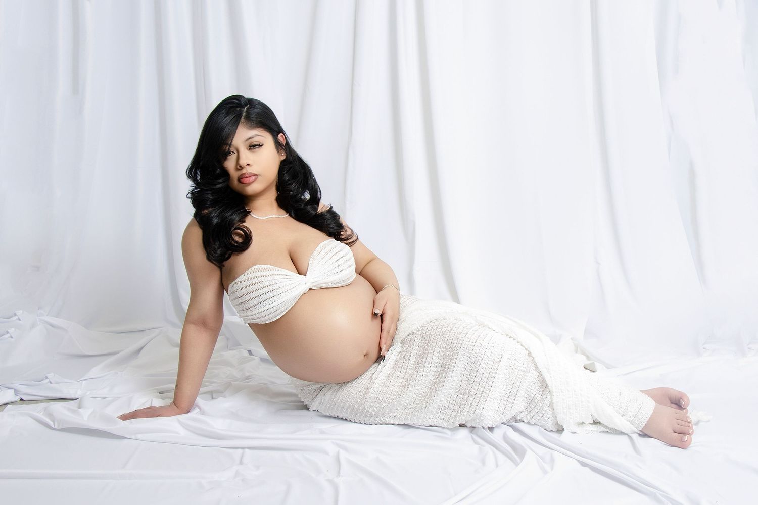 Elegant maternity session with subject reclining in white against a light backdrop.