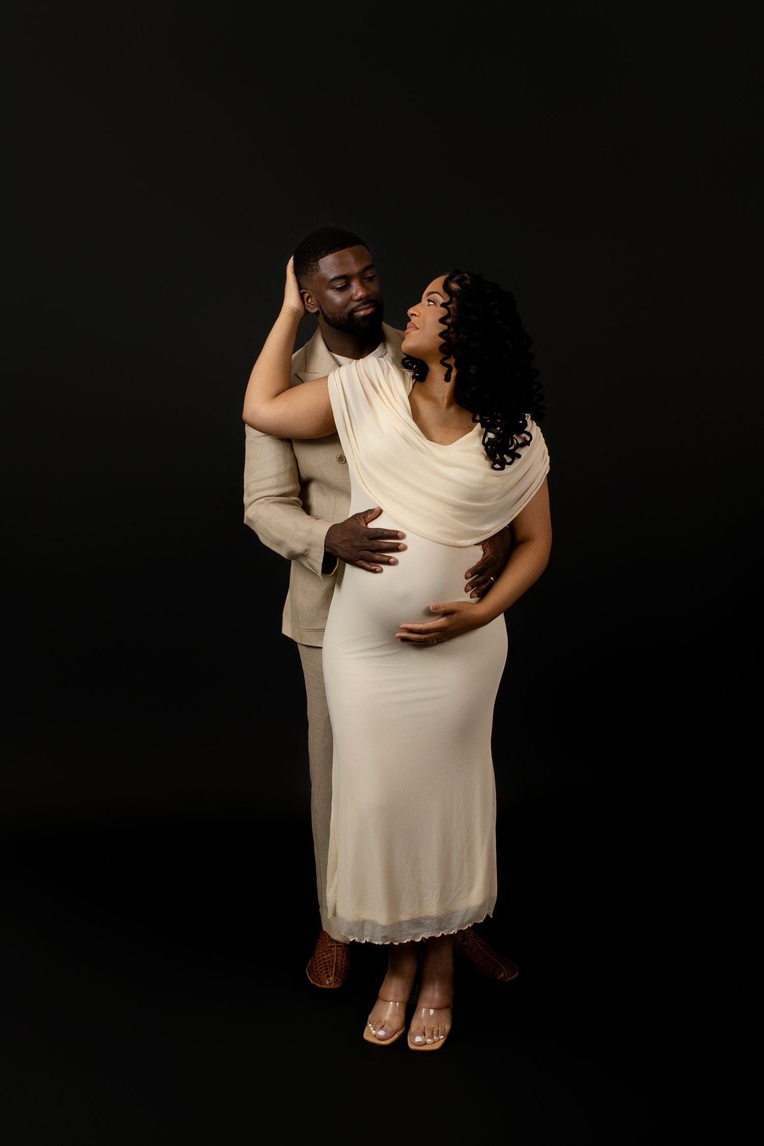 Intimate maternity studio portraits with couple in white and cream clothing against dark backdrop.